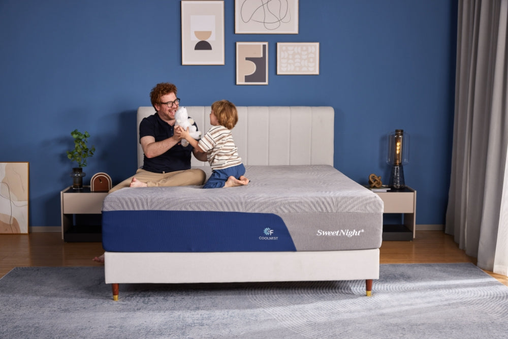 A father and his young son happily playing with a stuffed animal on a SweetNight Prime memory foam mattress in a stylish, modern blue bedroom.
