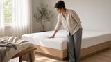 Morning view of a stripped white mattress with a shallow body-shaped dip in the sleep zone, a person pausing while making the bed