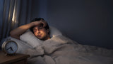 Tired adult lying awake in bed at dawn beside an analog alarm clock, illustrating sleep debt and lost sleep